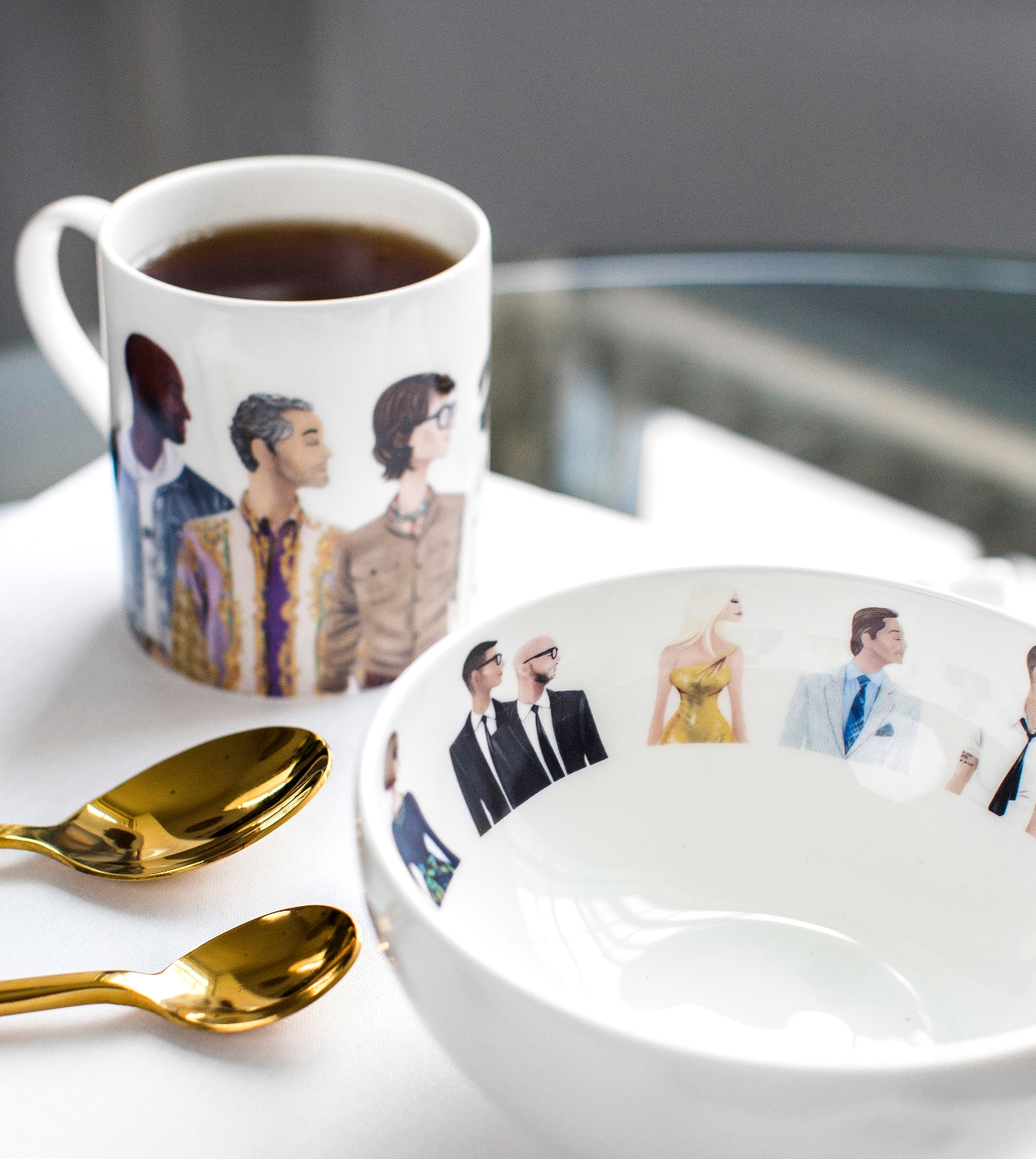 white bowl with famous people on the inside rim in cartoon form also a matching mug and gold spoon on table